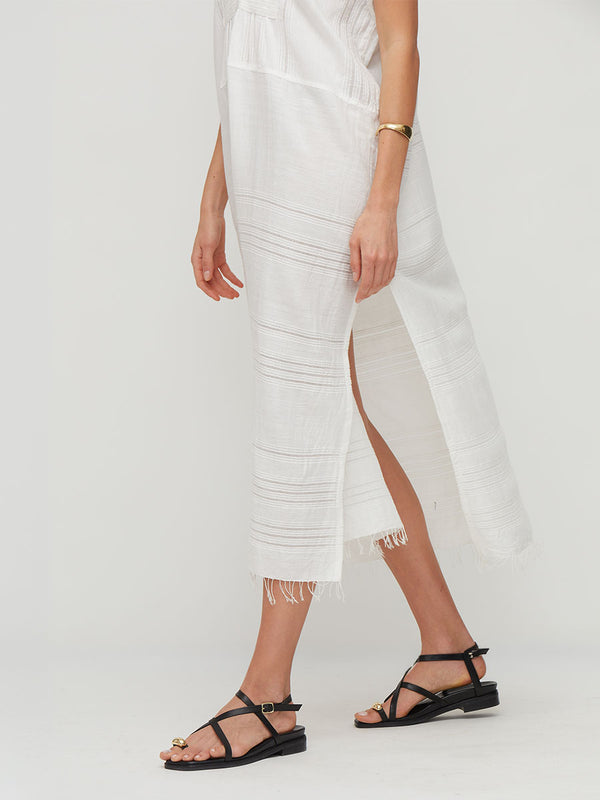 A model wearing a white lemlem handwoven GASIRA Caftan featuring  side slit and frayed hem, paired with black strappy flat sandals against a light gray studio background.