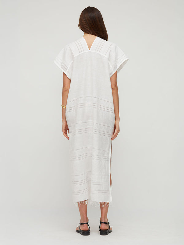 A woman viewed from the back wearing a lemlem handwoven GASIRA loose white maxi dress with short sleeves, a split V-neckline, and subtle horizontal stitched details, paired with black sandals against a light gray studio background.