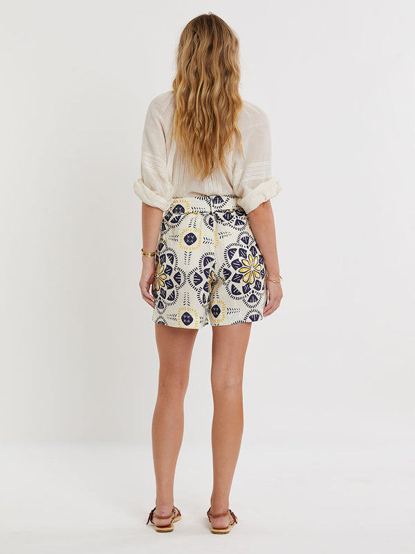 Woman standing wearing lemlem linen FAIZAH Shorts featuring floral inspired pattern and handwoven MITA Blouse in sheer fabric in cream color
