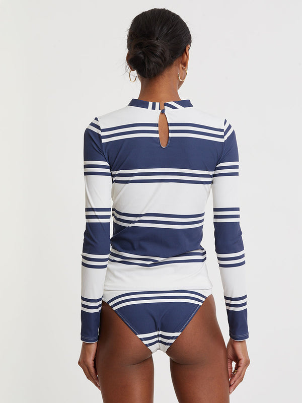 Woman standing wearing lemlem AZEB Rashguard and MERON Brief Bottom featuring bold two-tone stripes in white and classic navy, printed on soft ribbed fabric.