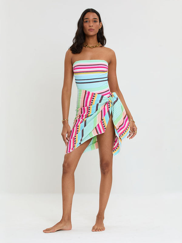 Woman Standing Wearing lemlem handwoven LEMA Sarong and One Piece Swimsuit  featuring intricate stripes and a tibeb pattern in vivid combinations of green, aqua, turquoise, pink, and soft peach-tangerine tones.