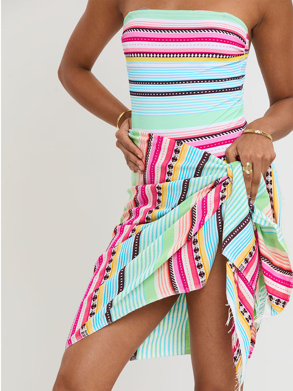Woman Standing Wearing lemlem handwoven LEMA Sarong and One Piece Swimsuit  featuring intricate stripes and a tibeb pattern in vivid combinations of green, aqua, turquoise, pink, and soft peach-tangerine tones.