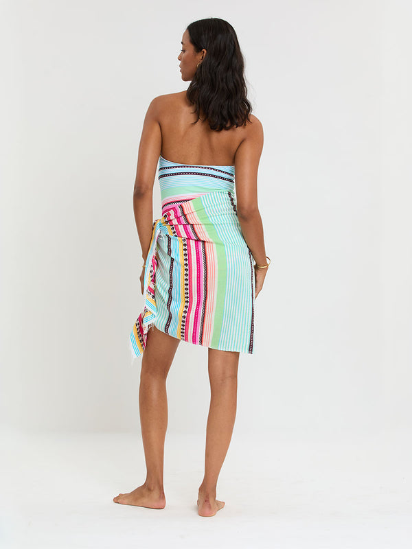 Woman Standing Wearing lemlem handwoven LEMA Sarong and One Piece Swimsuit  featuring intricate stripes and a tibeb pattern in vivid combinations of green, aqua, turquoise, pink, and soft peach-tangerine tones.