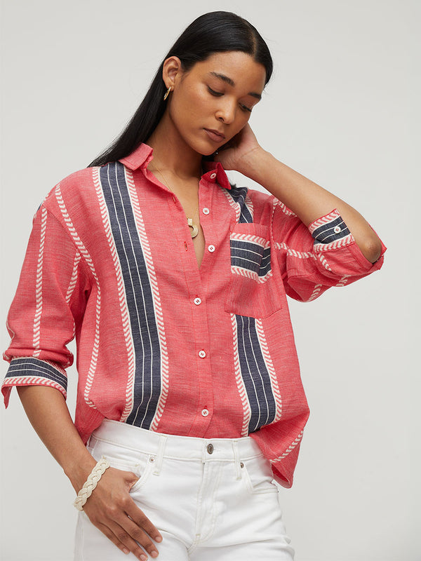 A woman wearing a coral pink linen button-up shirt with vertical navy and white striped details, paired with white jeans, against a light gray studio background.