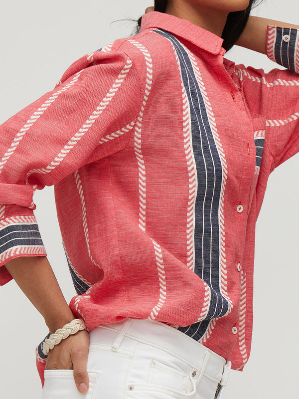 A close-up of a woman wearing a coral pink linen button-up shirt with vertical navy and white striped details, paired with white jeans.