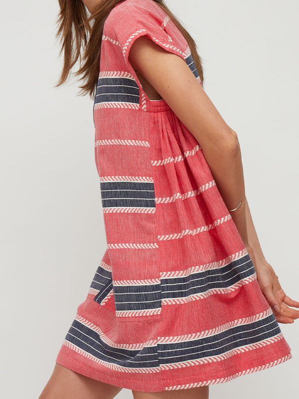 A woman wearing a coral pink linen dress with navy and white horizontal stripes, shown from a side-back angle against a light gray studio background.