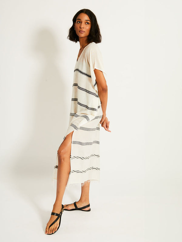 Side of a Woman Standing Wearing lemlem DALILA Caftan featuring  black intricate stripes and a Tibeb wave band on a soft vanilla ground