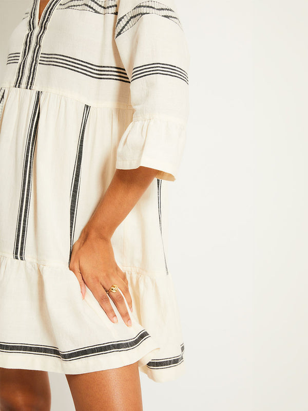 Close up on a Woman Standing Wearing lemlem HANNA Flutter Dress featuring  black intricate stripes and a Tibeb wave band on a soft vanilla ground