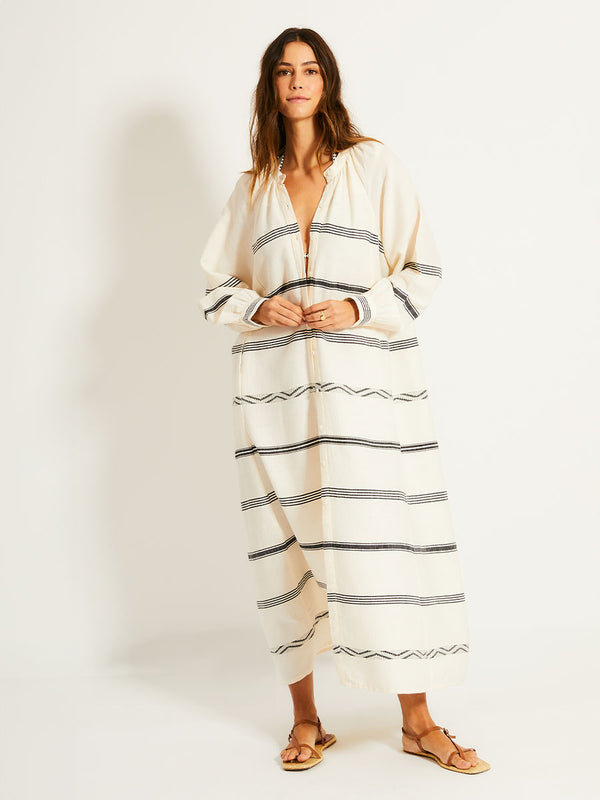 Woman Standing Wearing lemlem MAKEDA Dress featuring  black intricate stripes and a Tibeb wave band on a soft vanilla ground