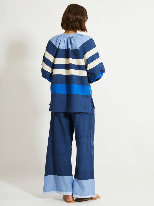 Back shot of a Woman Standing Wearing lemlem MITA Blouse featuring a striking color-block combination of three shades of blue, paired with bands of neutral cream and white