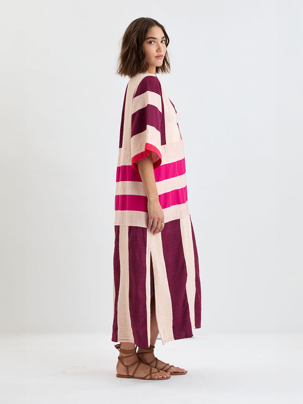 Side shot of a Woman standing wearing lemlem EDNA Dress  featuring a striking color-block combination of vivid magenta, scarlet, and burgundy colors.