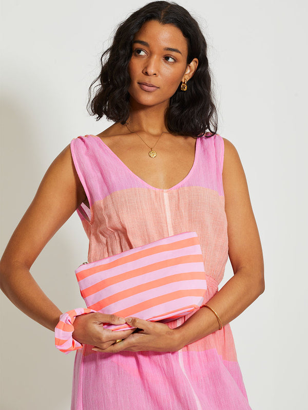 Woman Standing Wearing lemlem Melat Scoop Dress featuring gauzy fabric with a beautiful ombre color block of pink, blush, and peach colors, holding pouch and a scrunchie  featuring stripe pattern in orange and pink colors