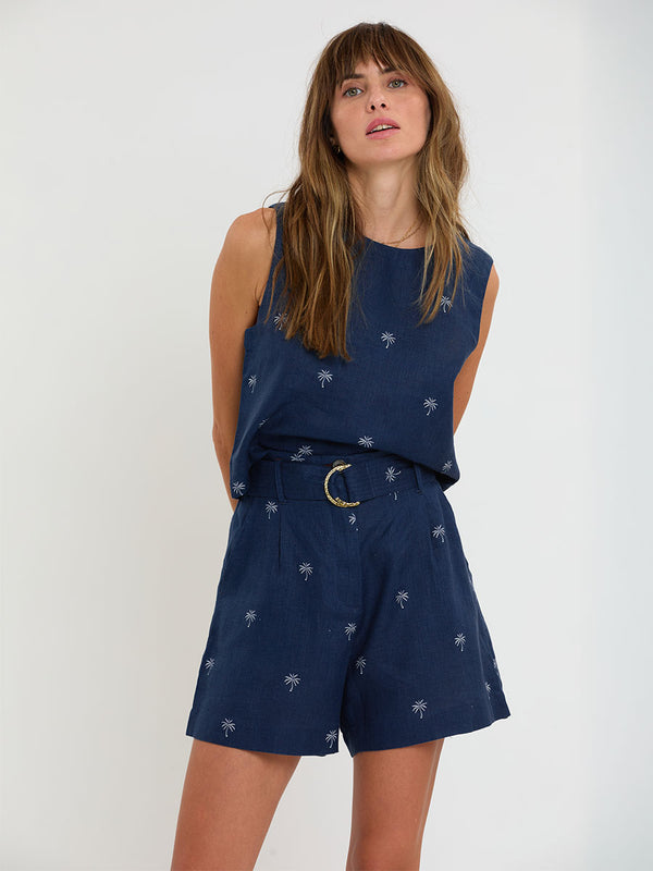 Woman Standing Wearing lemlem ADOWA Shell Tank and FAIZAH Shorts featuring  ivory palm tree motifs set against a rich navy background.