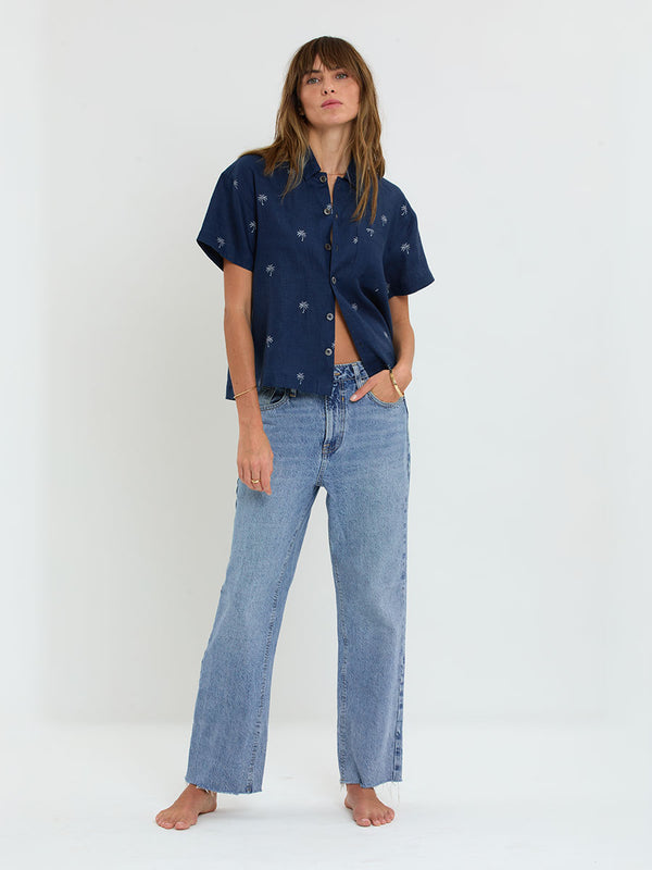 Woman Standing Wearing lemlem TUULI Cabana Shirt featuring  ivory palm tree motifs set against a rich navy background.