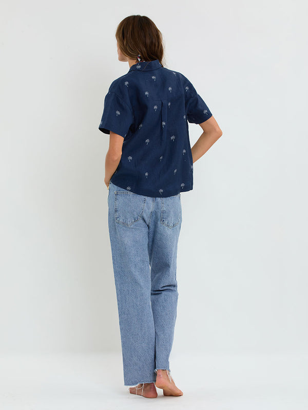 Back of a Woman Standing Wearing lemlem TUULI Cabana Shirt featuring  ivory palm tree motifs set against a rich navy background.