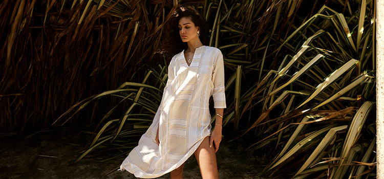 Woman standing in front of palm bushes wearing a lemlem hand woven Fana maxi caftan dress in cream with wide white blocks.