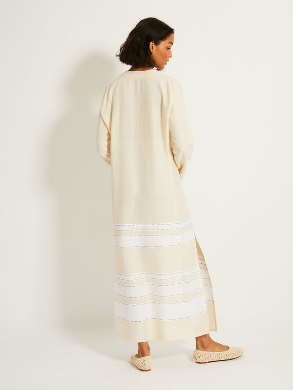Back of a Woman Standing Wearing lemlem THEODORA Dress featuring bold stripes in bright white on a vanilla cream background, accented with navy pick stitch detailing