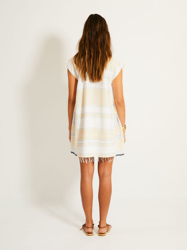 Back of a Woman Standing Wearing lemlem ELINA Caftan featuring bold stripes in bright white on a vanilla cream background, accented with navy pick stitch detailing
