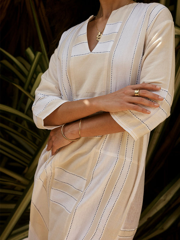 Close up on a Woman Standing Wearing lemlem FANA Caftan featuring  bold stripes in bright white on a vanilla cream background, accented with navy pick stitch detailing