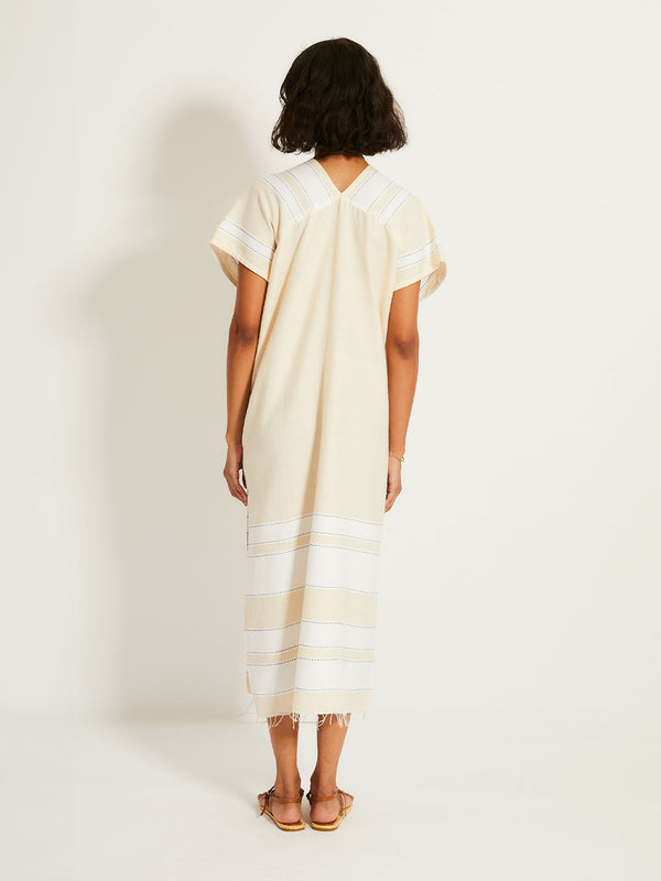 Back of a Woman Standing Wearing lemlem GASIRA Caftan featuring bold stripes in bright white on a vanilla cream background, accented with navy pick stitch detailing