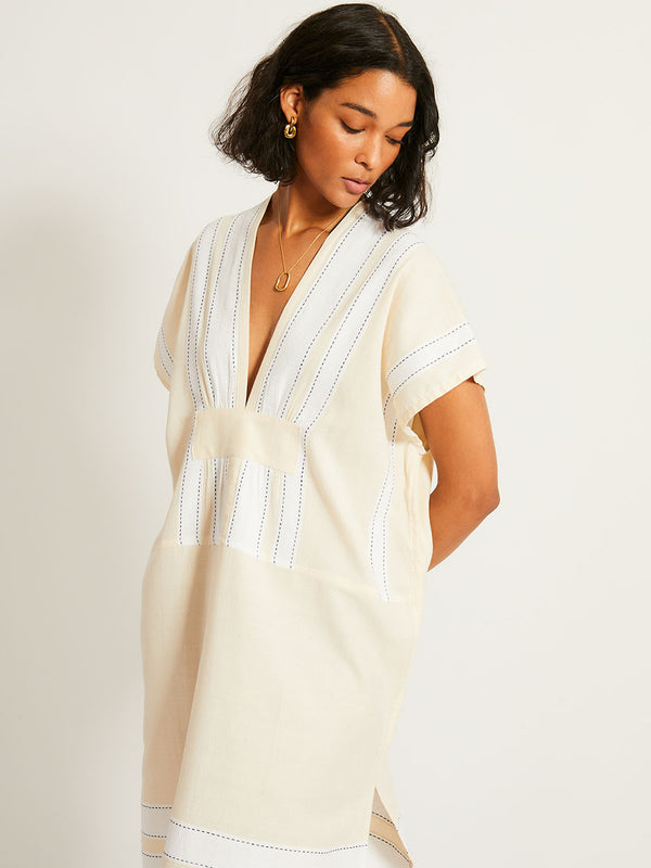 Close up on a Woman Standing Wearing lemlem GASIRA Caftan featuring bold stripes in bright white on a vanilla cream background, accented with navy pick stitch detailing