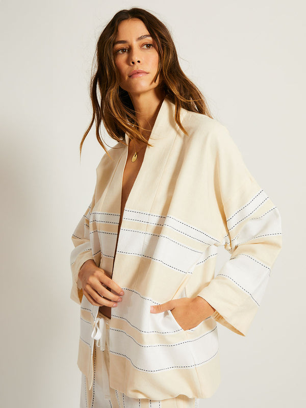 Woman Standing Wearing lemlem TERARA Jacket featuring bold stripes in bright white on a vanilla cream background, accented with navy pick stitch detailing