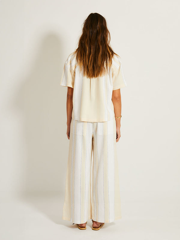Back of a Woman Standing Wearing lemlem TUULI Shirt featuring bold stripes in bright white on a vanilla cream background, accented with navy pick stitch detailing