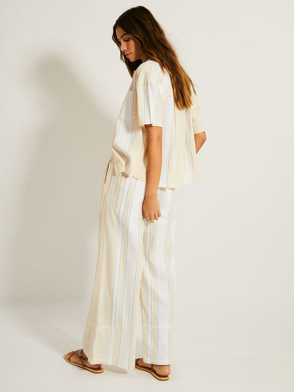Side Shot of a Woman Standing Wearing lemlem TUULI Shirt featuring bold stripes in bright white on a vanilla cream background, accented with navy pick stitch detailing