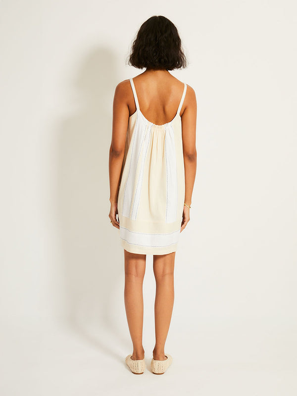 Back of a Woman Standing Wearing lemlem ZINA Dress featuring bold stripes in bright white on a vanilla cream background, accented with navy pick stitch detailing