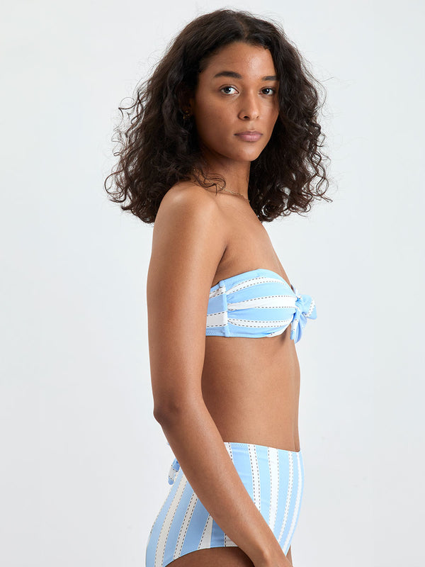Side shot of a Woman wearing  lemlem ELSI High Waist bottom and AVA Bandeau Top featuring bold, crisp white stripes on a sky-blue background, accented with black detailing.