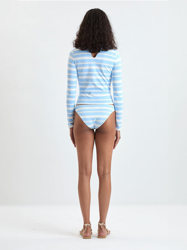 Woman wearing  lemlem AZEB Rash Guard and MERON Birief Bottom featuring bold, crisp white stripes on a sky-blue background, accented with black detailing.
