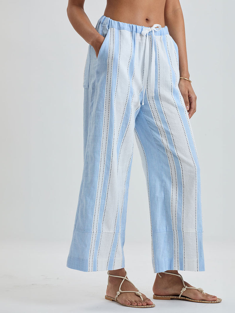 Woman Standing wearing lemlem DESTA Pants featuring bold, crisp white stripes on a sky-blue background, accented with black detailing.