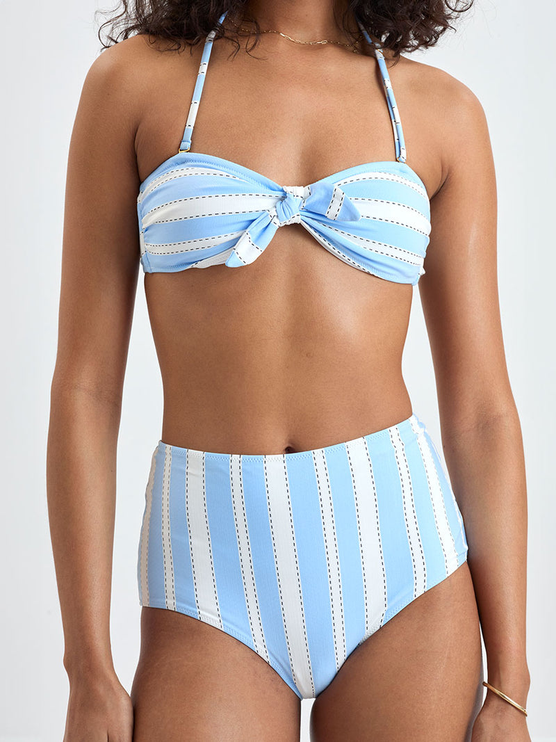 Close up on a Woman wearing  lemlem ELSI High Waist bottom and AVA Bandeau Top featuring bold, crisp white stripes on a sky-blue background, accented with black detailing.