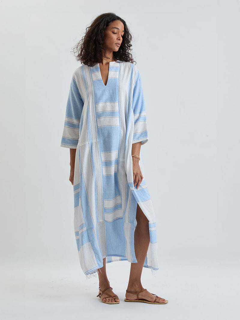 Woman wearing a lemlem  handwoven FANA Caftan  featuring bold, crisp white stripes on a sky-blue background, accented with black stitch detailing