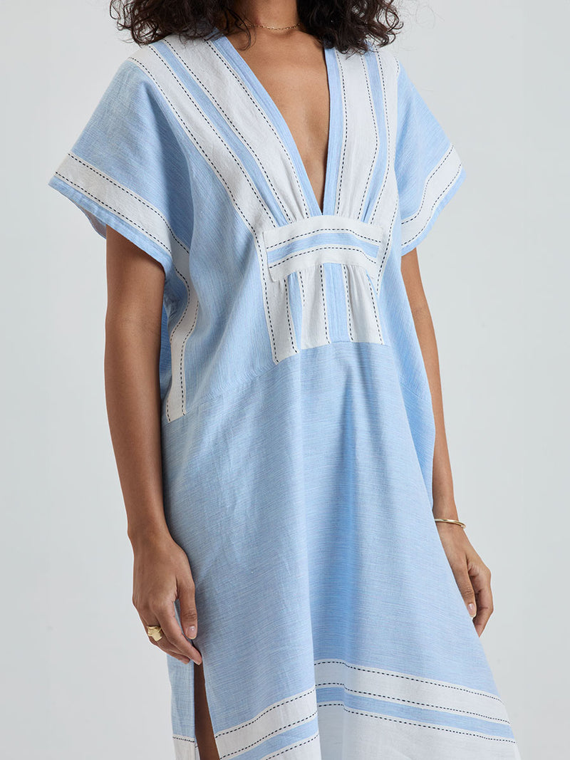 Close up on a woman wearing lemlem GASIRA Caftan featuring color block stripe pattern in white on sky blue background