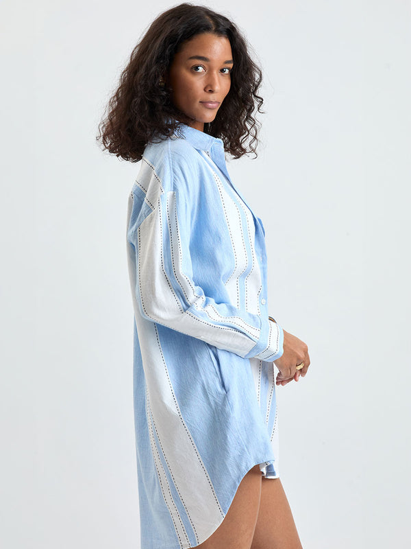 Side shot of a Woman Standing wearing lemlem MARIAM Handwoven shirt featuring bold, crisp white stripes on a sky-blue background, accented with black stitch detailing