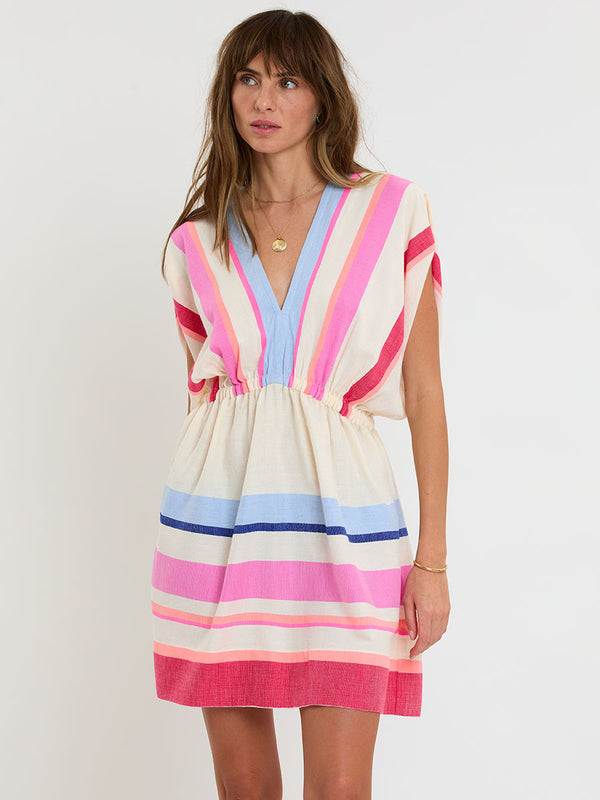 Woman standing wearing lemlem handwoven ALEM Plunge Dress featuring bold color block stripe pattern in pink, peach, red, sky and marine colors on vanilla background.