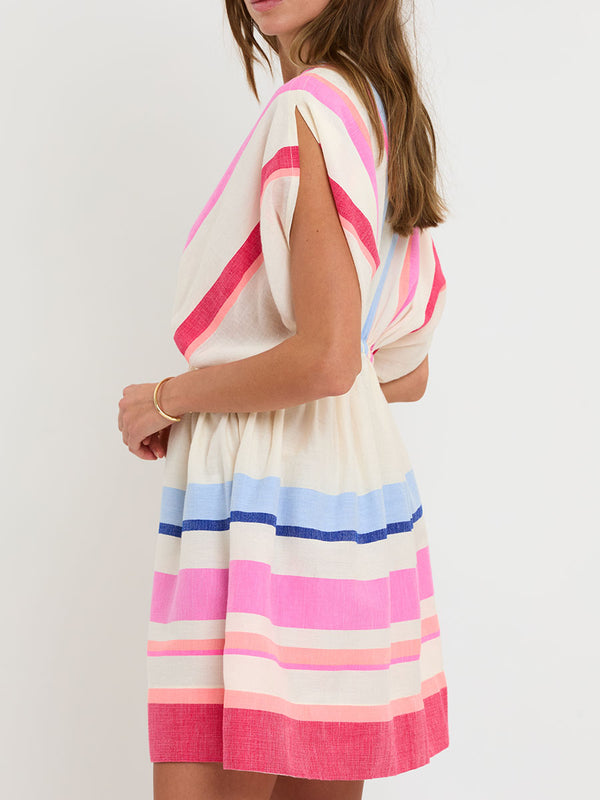 Woman standing wearing lemlem handwoven ALEM Plunge Dress featuring bold color block stripe pattern in pink, peach, red, sky and marine colors on vanilla background.