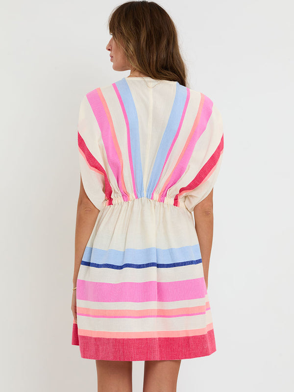 Woman standing wearing lemlem handwoven ALEM Plunge Dress featuring bold color block stripe pattern in pink, peach, red, sky and marine colors on vanilla background.