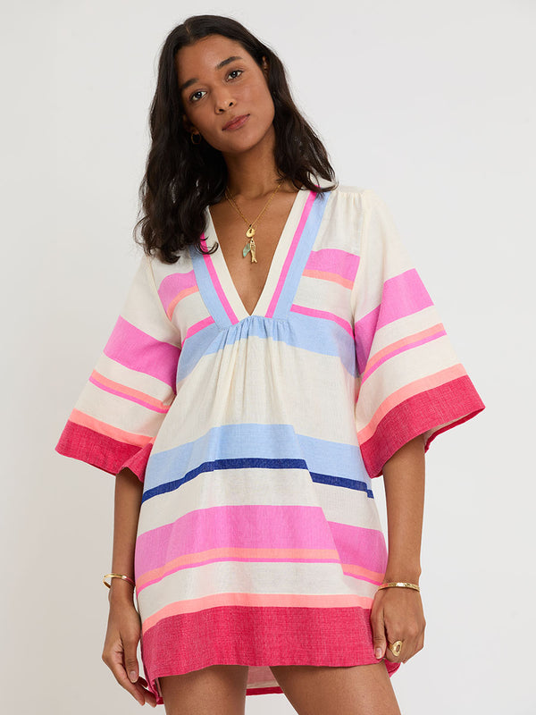 Woman standing wearing lemlem handwoven BELKIS Caftan featuring bold color block stripe pattern in pink, peach, red, sky and marine colors on vanilla background.