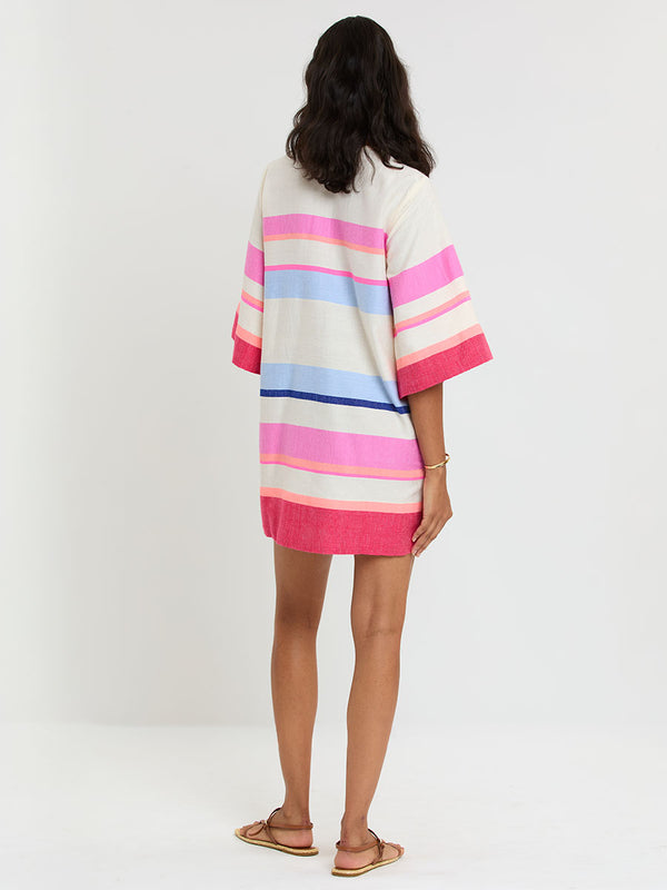 Woman standing wearing lemlem handwoven BELKIS Caftan featuring bold color block stripe pattern in pink, peach, red, sky and marine colors on vanilla background.
