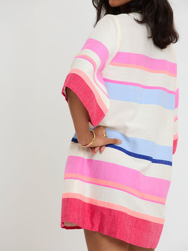 Woman standing wearing lemlem handwoven BELKIS Caftan featuring bold color block stripe pattern in pink, peach, red, sky and marine colors on vanilla background.