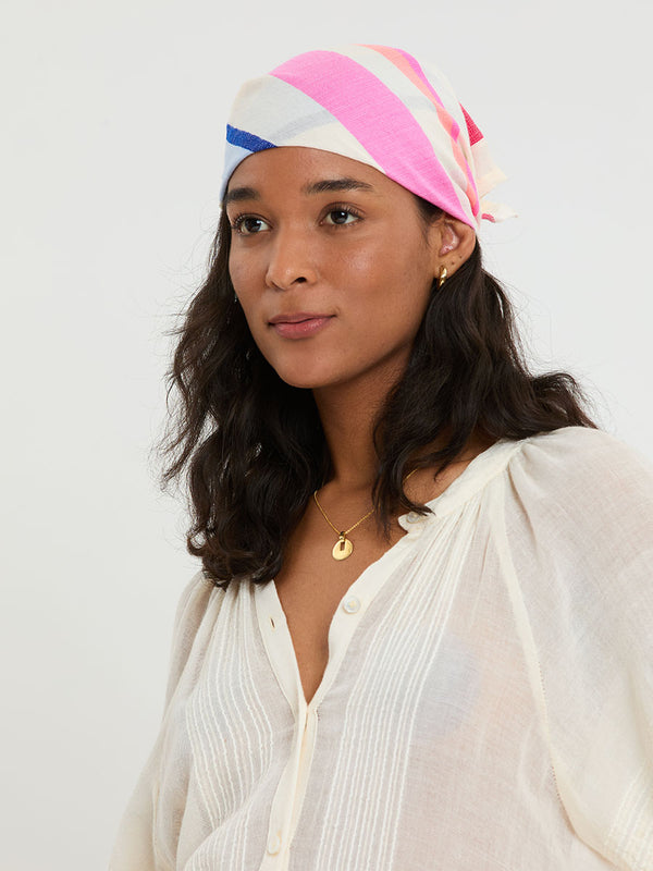  Woman standing wearing lemlem handwoven DANA Bandana featuring bold color block stripe pattern in pink, peach, red, sky and marine colors on vanilla background and blue jeans