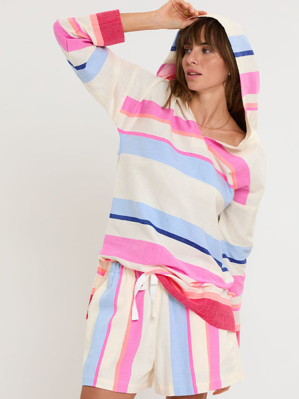 Woman standing wearing lemlem handwoven HENOK Hoodie and SAFIA Shorts featuring bold color block stripe pattern in pink, peach, red, sky and marine colors on vanilla background.