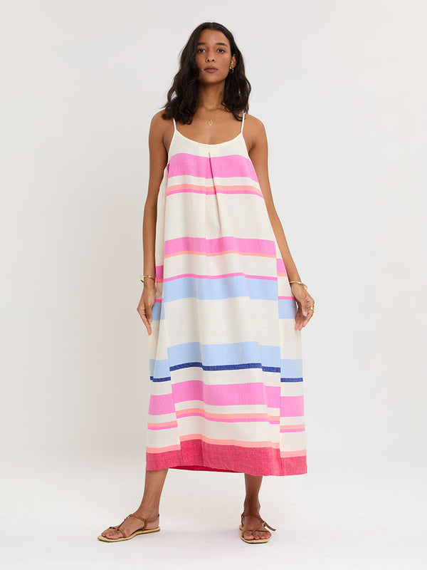 Woman standing wearing lemlem handwoven NIA Slip Dress featuring bold color block stripe pattern in pink, peach, red, sky and marine colors on vanilla background.