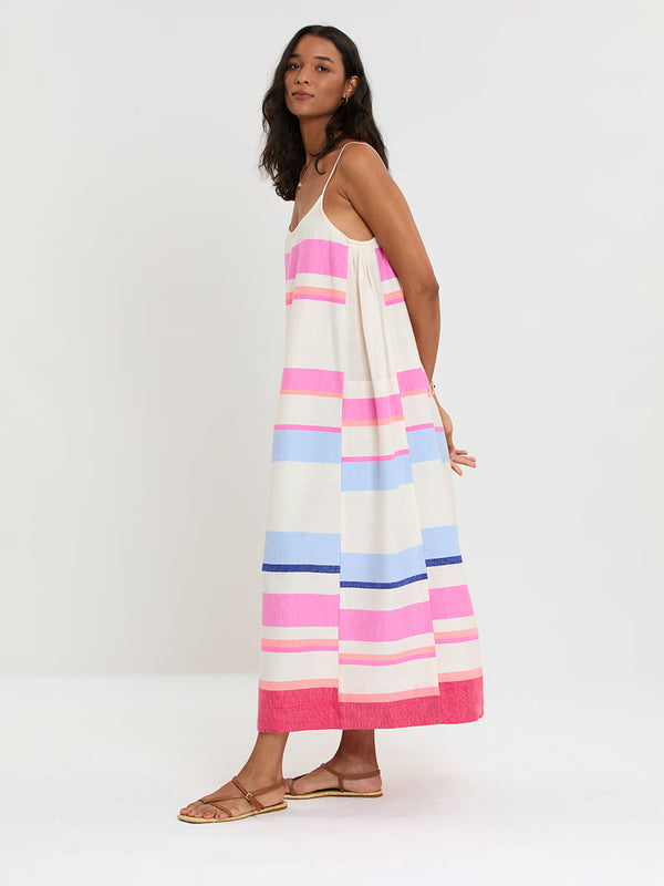 Woman standing wearing lemlem handwoven NIA Slip Dress featuring bold color block stripe pattern in pink, peach, red, sky and marine colors on vanilla background.