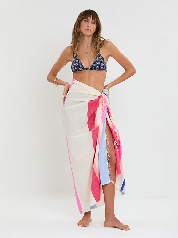 Woman standing wearing lemlem handwoven TALAKI Sarong featuring bold color block stripe pattern in pink, peach, red, sky and marine colors on vanilla background.