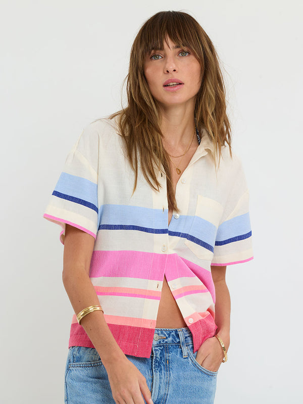 Woman standing wearing lemlem handwoven TUULI Cabana Shirt featuring bold color block stripe pattern in pink, peach, red, sky and marine colors on vanilla background and blue jeans