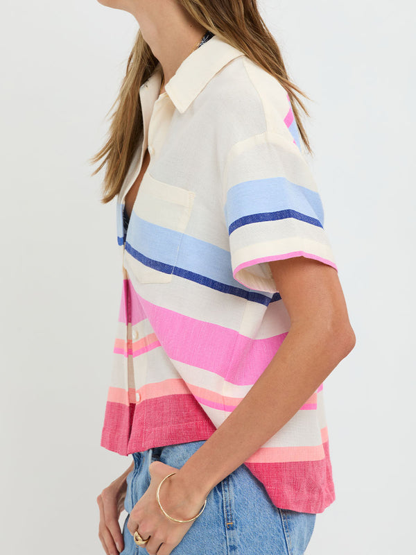 Woman standing wearing lemlem handwoven TUULI Cabana Shirt featuring bold color block stripe pattern in pink, peach, red, sky and marine colors on vanilla background and blue jeans