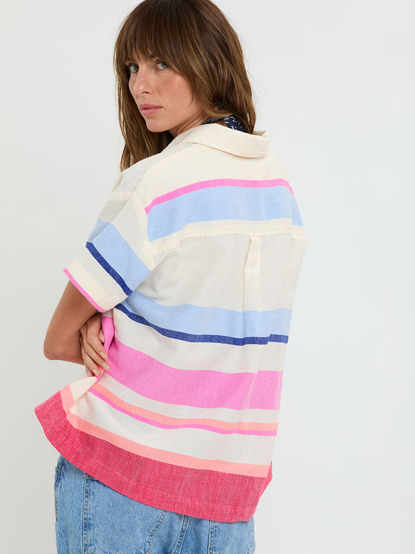 Woman standing wearing lemlem handwoven TUULI Cabana Shirt featuring bold color block stripe pattern in pink, peach, red, sky and marine colors on vanilla background and blue jeans